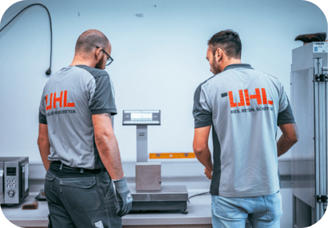 Zwei Techniker in UHL-Arbeitshemden führen eine Qualitätskontrolle durch und wiegen eine Betonprobe.