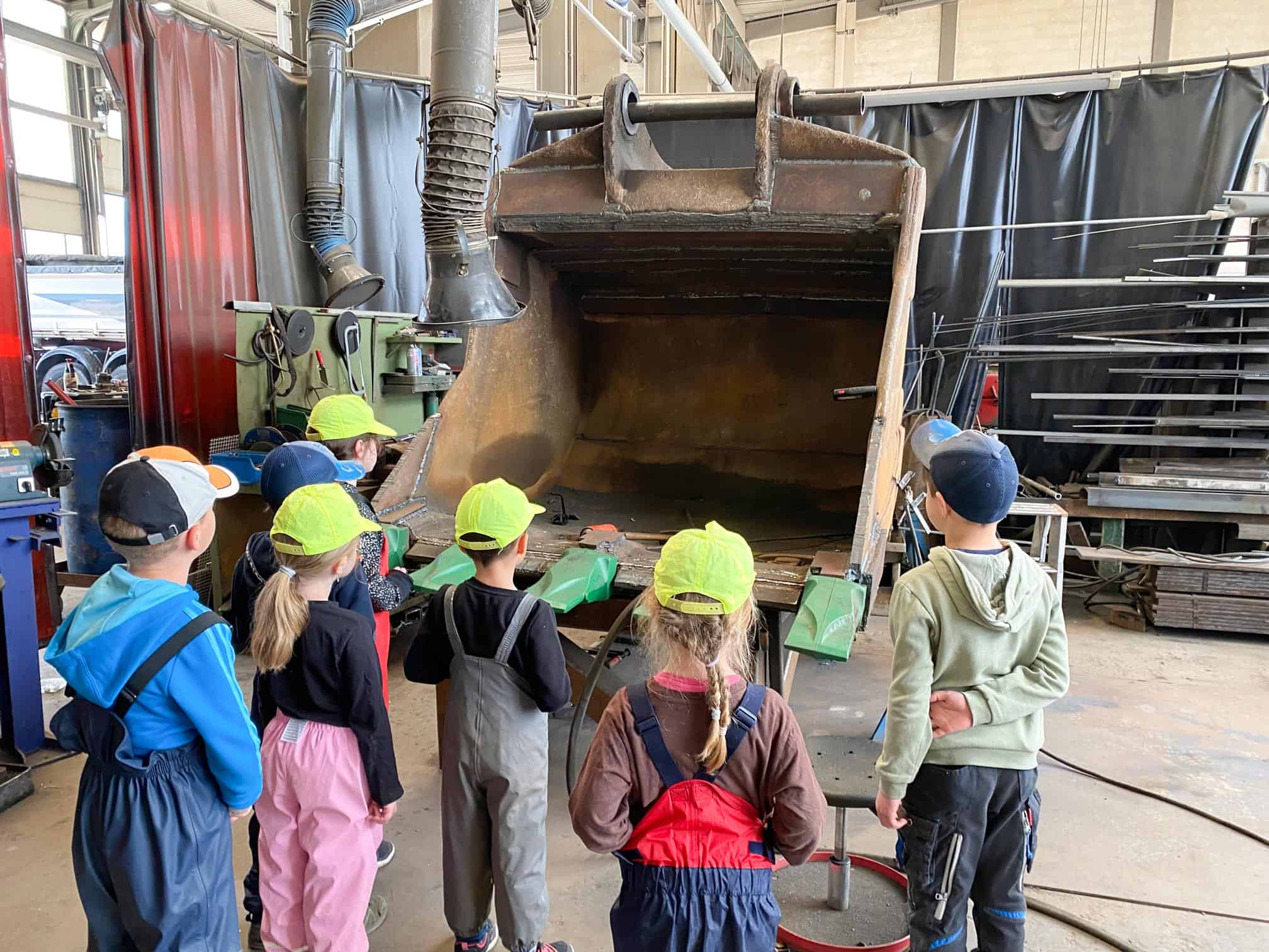 Kinder in Schutzausrüstung beobachten in einer Werkstatt eine große Baggerschaufel.