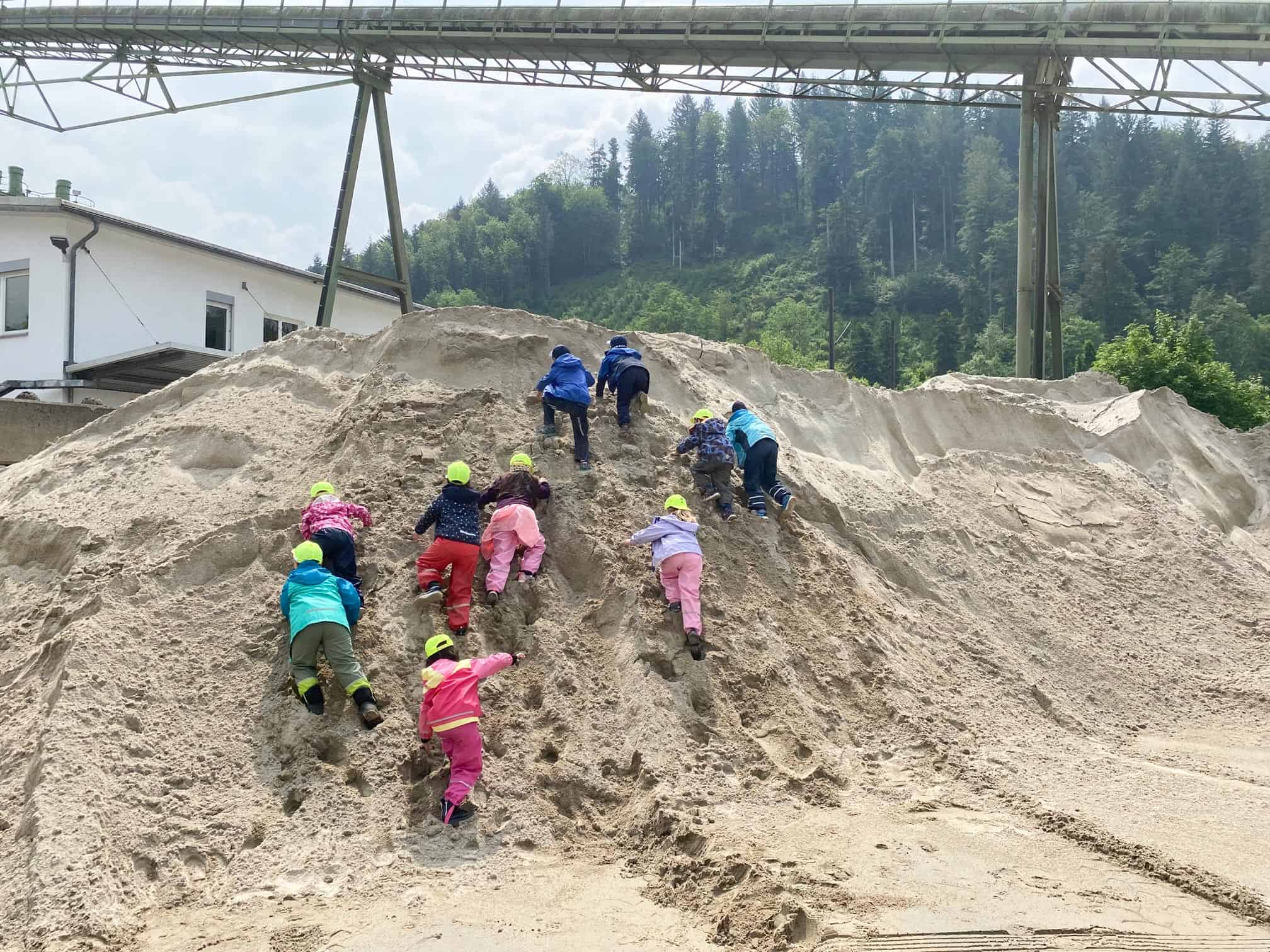 Kinder klettern mit Schutzhelmen auf einen großen Sandhaufen.