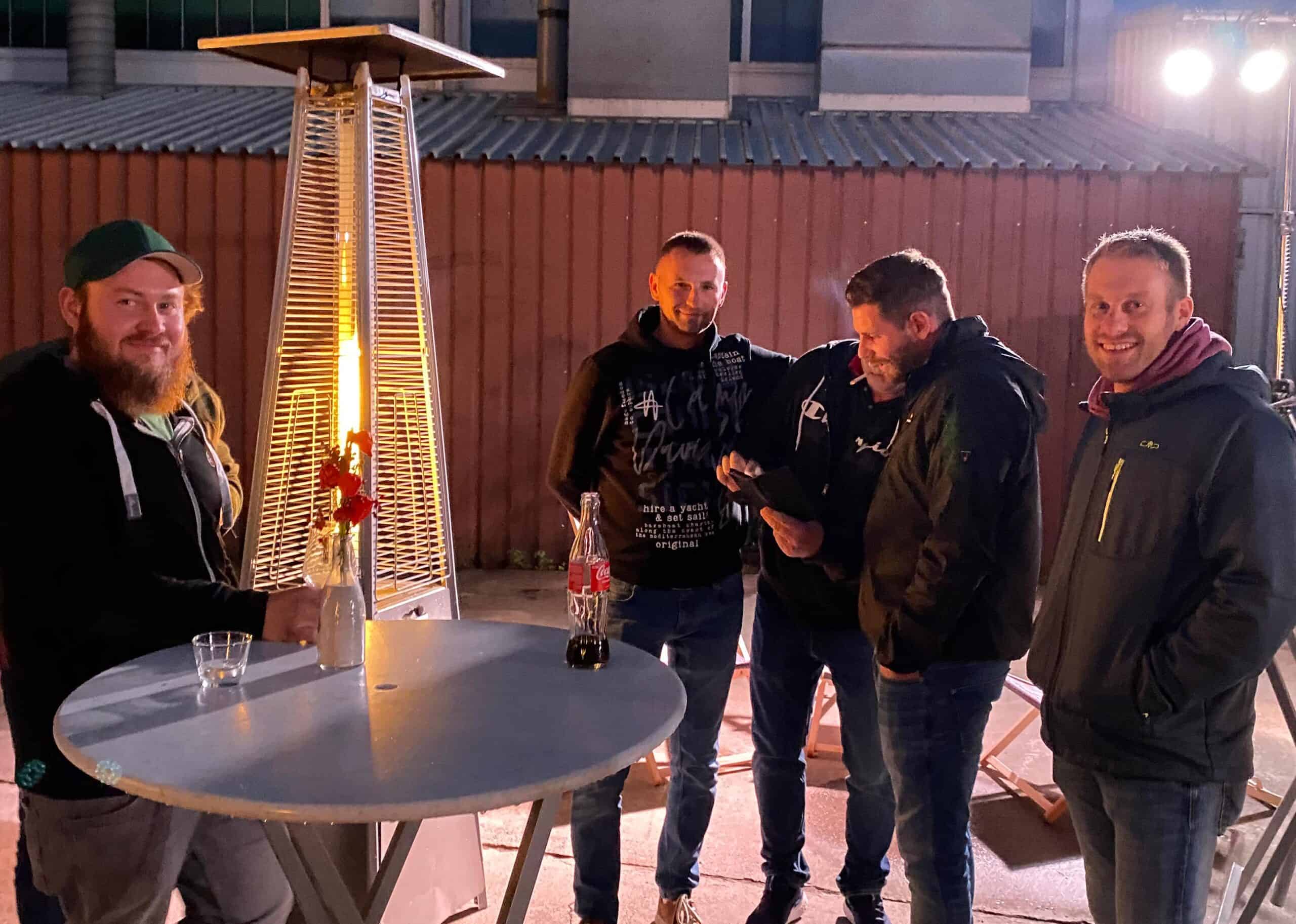 Vier Männer und eine Frau genießen Getränke und Gespräche im Freien in der Nähe eines Terrassenheizers.