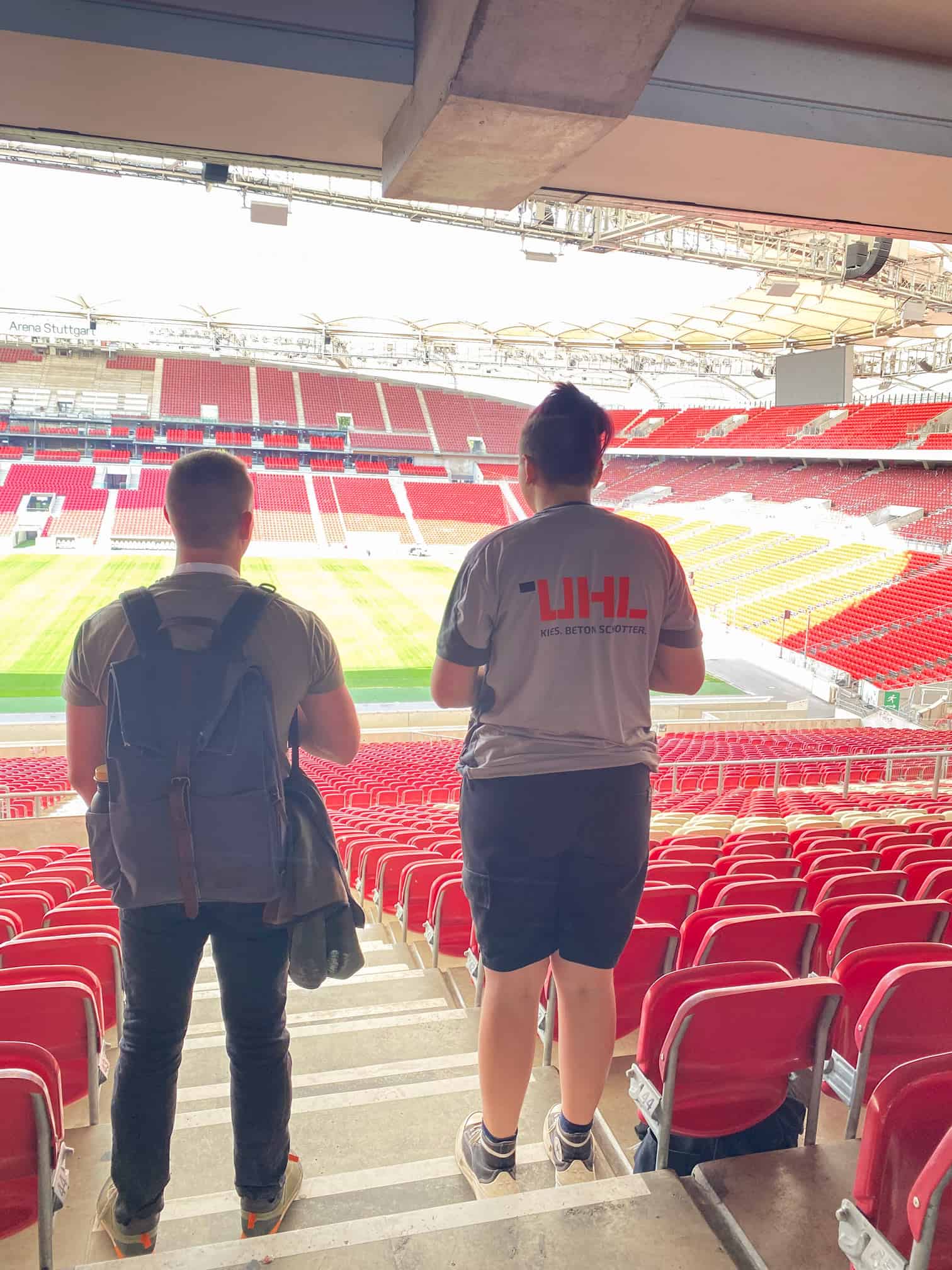 Zwei Personen stehen auf leeren Stadionsitzen und schauen auf das Spielfeld. Eine trägt ein UHL-Trikot von Kies, Beton, Schotter.
