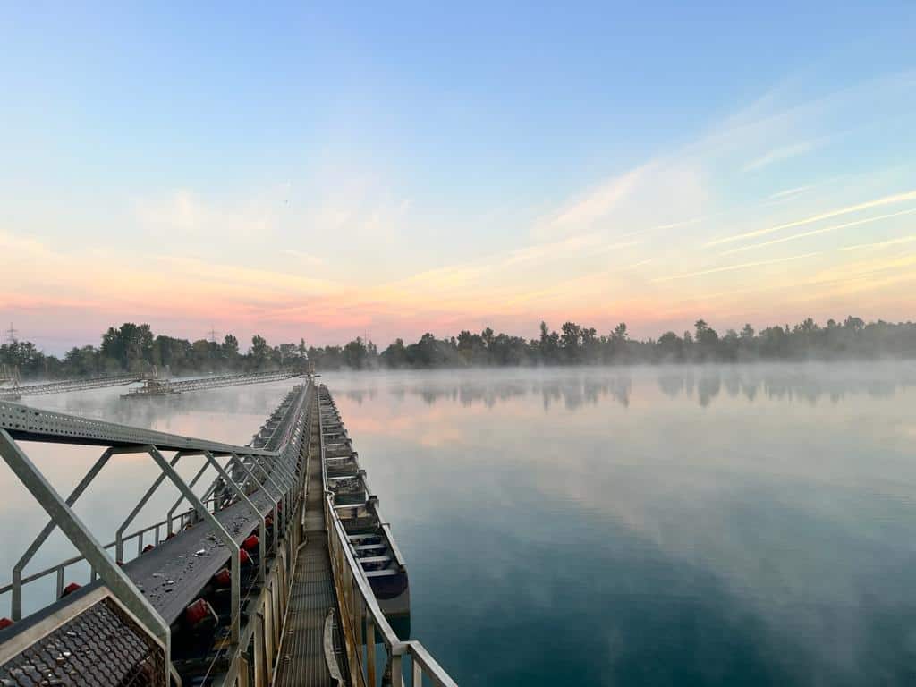 Sonnenaufgang über ruhigem Wasser mit einem langen Förderbandsystem, das sich zum Ufer erstreckt.
