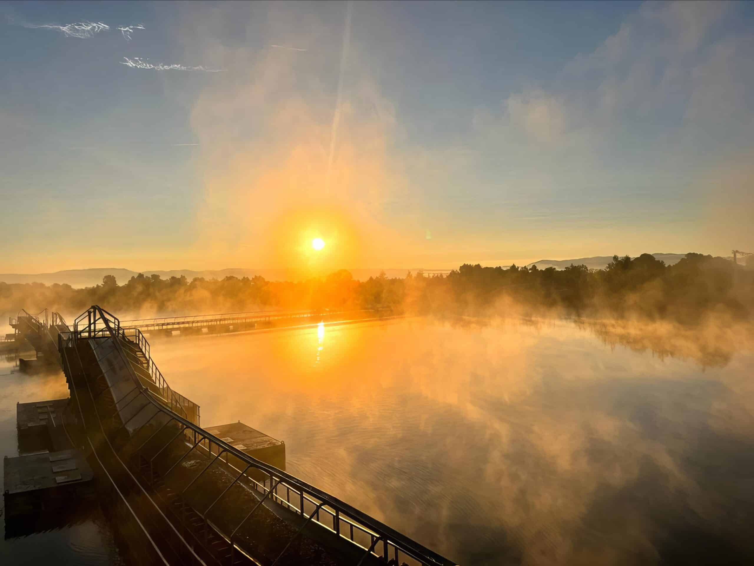 Sonnenaufgang über ruhigem Wasser mit Nebel, der goldenes Licht reflektiert. Industrielle Infrastruktur sichtbar.